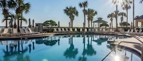 A clear swimming pool surrounded by palm trees and lounge chairs, with a gazebo in the background.