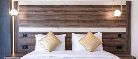 Una cama bien hecha con sábanas blancas y dos almohadones decorativos, un cabecero de madera con una lámpara, y una mesita de noche con toma de corriente.