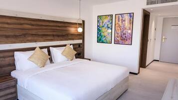 Een moderne hotelkamer met een houten hoofdeinde, een groot bed met witte lakens, twee abstracte schilderijen aan de muur en een deur rechts.