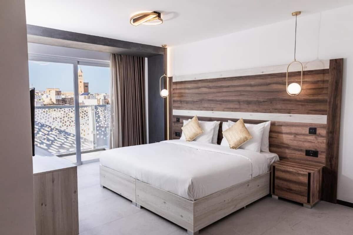 A modern bedroom with a large bed, wooden headboard, and a city view through large windows.