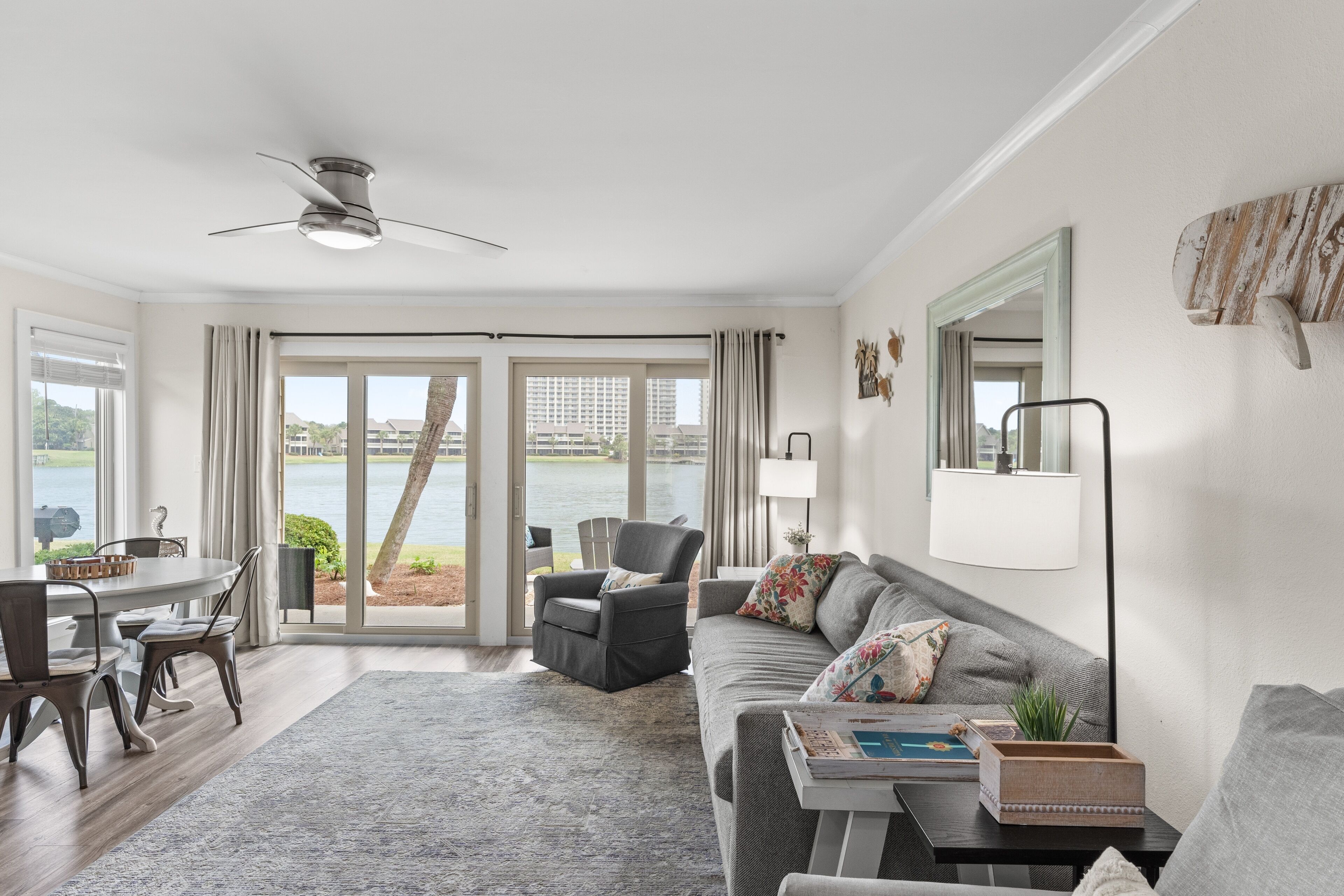 A modern living room with a dining area, a ceiling fan, a sofa, a coffee table, and a view of a body of water through large glass doors.