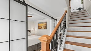 Un interior moderno con una escalera de madera, una cama con cabecera blanca y un cuadro en la pared.