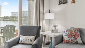 Una sala moderna con un sillón gris, una mesita blanca y un sofá con un cojín floral de colores. Un cojín que dice "Beach" está en el sillón, y un letrero que dice "Welcome to Paradise" está en la pared.