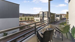 Un balcón con sillas de mimbre y una mesa de vidrio, con vistas a un área ajardinada y edificios con colinas al fondo.