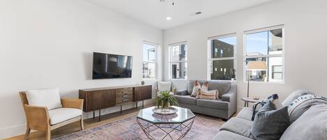 A spacious living room with a sofa, armchair, and a coffee table. There is a flat-screen TV mounted on the wall, a ceiling fan, and large windows providing natural light.