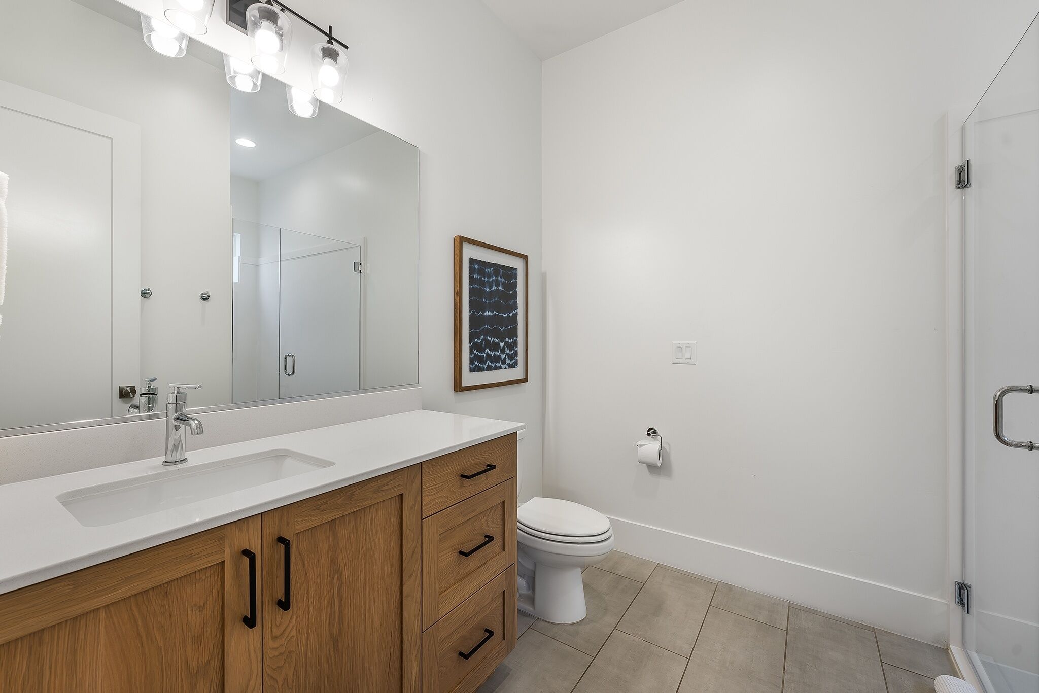 A modern bathroom with a white sink, wooden vanity, a large mirror, and a framed artwork on the wall.