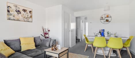 A modern living room with a grey sofa, yellow cushions, a small table, and a dining area with yellow chairs.