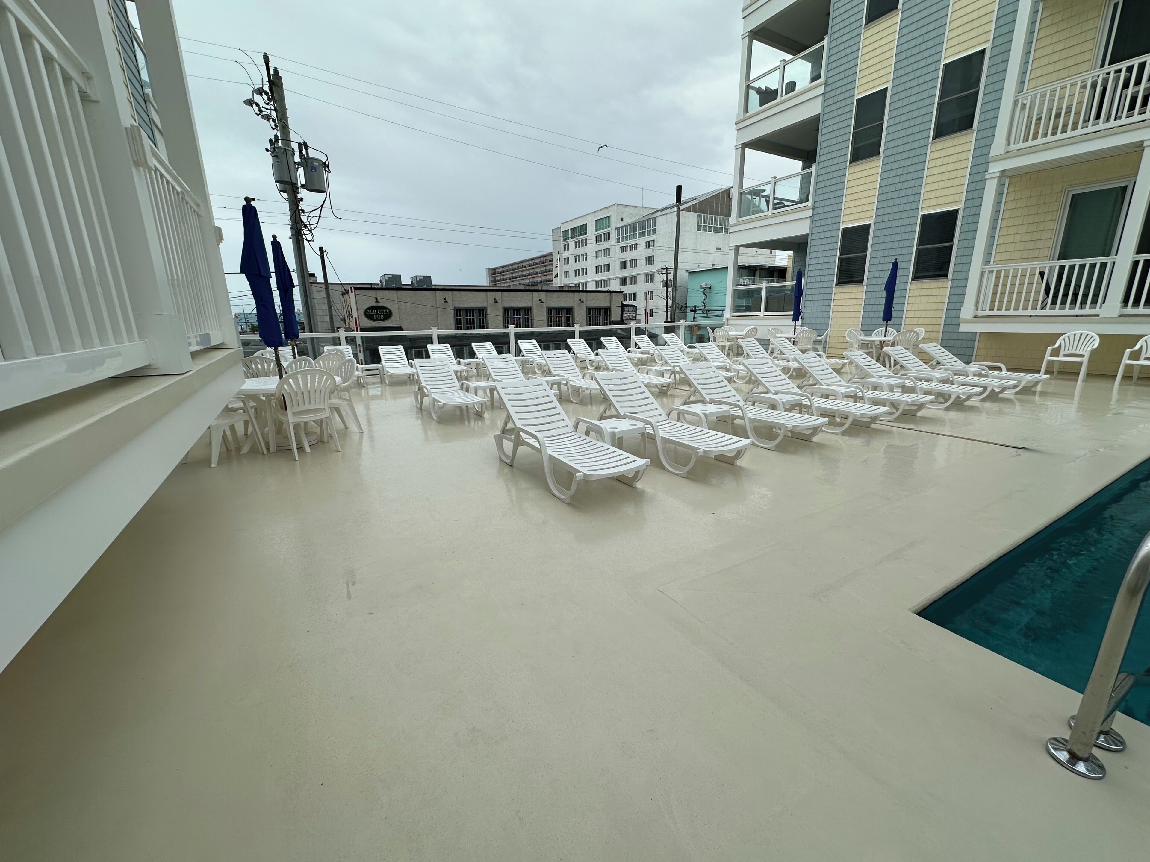 A poolside area with white lounge chairs arranged in rows, a pool, and a building with balconies in the background.