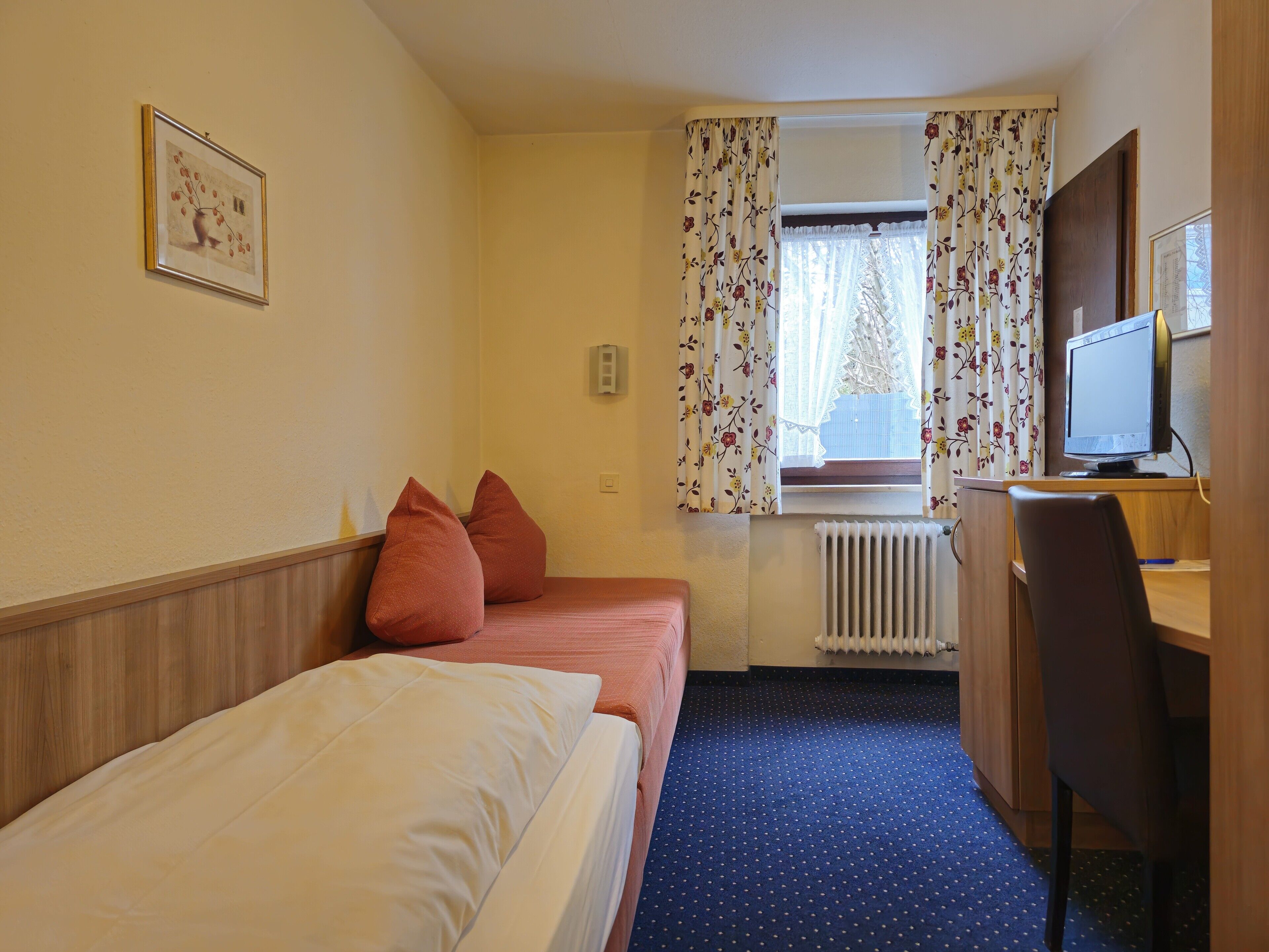 Habitación de hotel con dos camas individuales, un escritorio con computadora, un radiador y una ventana con cortinas florales.