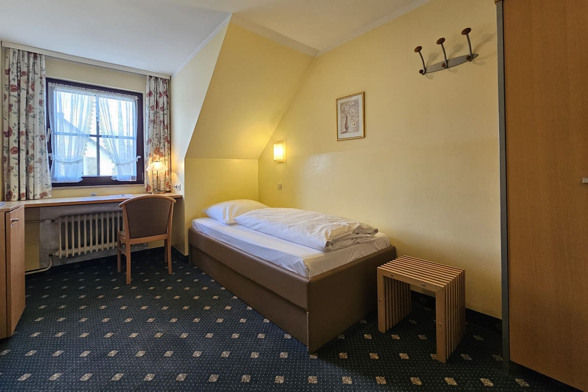 A compact room with a bed, a desk, a chair, a radiator, a window with curtains, a wall-mounted light, and a door.