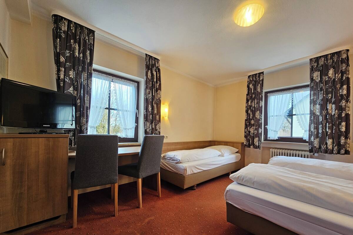 A hotel room with a bed, a desk, a chair, a television, and two windows with curtains.