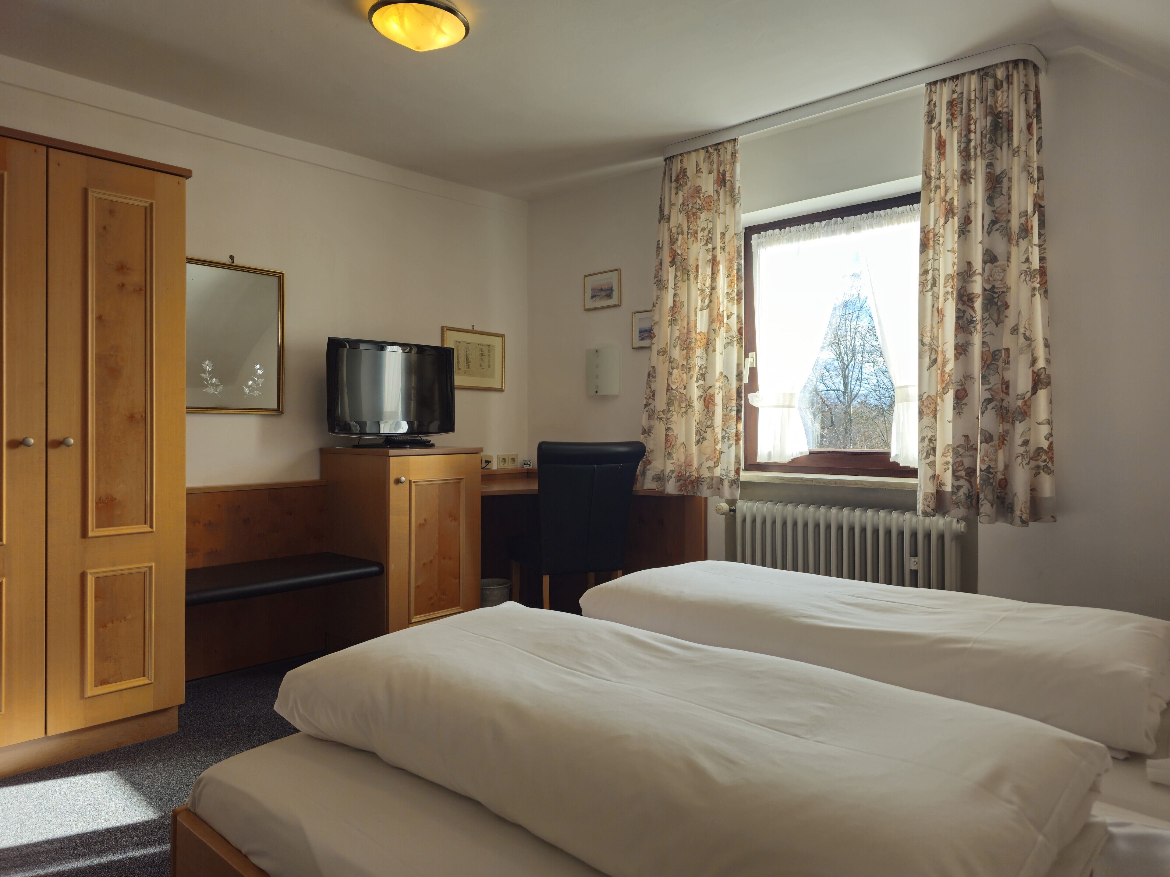 A hotel room with two beds, a desk with a television, a wardrobe, and a window with curtains.