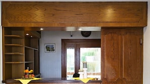 A wooden reception desk with a clock above it, a small shelf with fruits, and a view of a garden through a glass door. - ARRIVEL Hotel am Bergson (Munich)