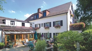 A two-story house with a covered patio area, a garden with potted plants, and a balcony with hanging flower baskets. - ARRIVEL Hotel am Bergson (Munich)