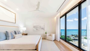 Ein modernes Schlafzimmer mit einem großen Bett, einem Nachttisch, einem Sessel und Meerblick durch große Fenster.