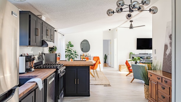 A modern kitchen with dark cabinets, a wooden countertop, and a central island. There is a dining area with orange chairs and a TV in the background.