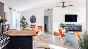 A modern kitchen with a wooden countertop, a stove, and a microwave. There is a dining area with orange chairs and a table. A television is mounted on the wall, and there are plants in the room.