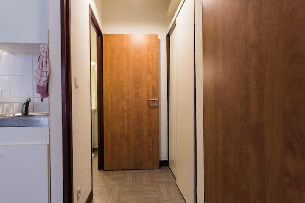Un couloir étroit, avec une porte en bois au fond, un mur recouvert de carreaux blancs et un petit comptoir surmonté d’un tissu.