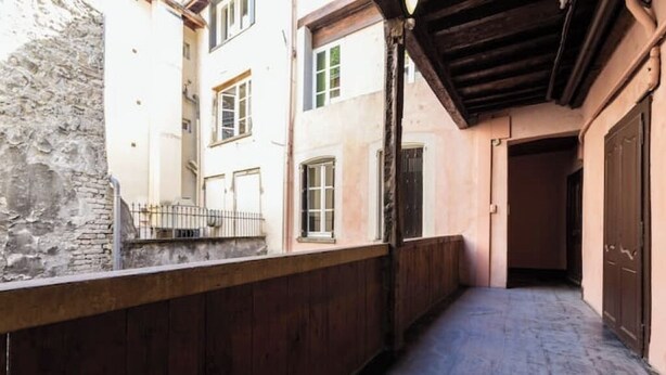 A narrow corridor with a wooden railing, a stone wall on one side, and a building entrance on the other. - Charming studio under the roof in Grenoble city center (Grenoble)