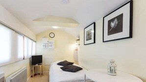 A small, well-lit room with a bed, a table, a clock, and framed pictures on the wall. - Charming studio under the roof in Grenoble city center (Grenoble)