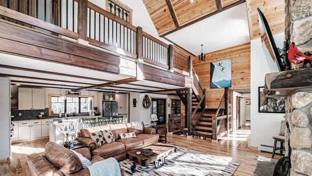 A spacious living area with a high ceiling, wooden beams, a large brown leather sofa, a dining table, and a kitchen in the background. - River access/Golf 2 miles/hiking/skiing/hot tub/sauna (Newry)