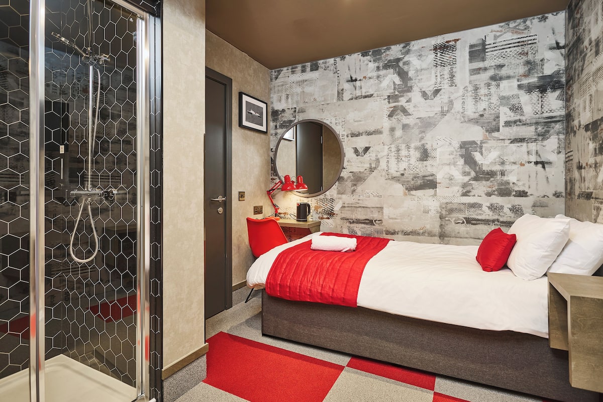 A hotel room with a shower, a bed with red and white bedding, a round mirror, and a wall with a textured pattern.