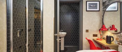 Une chambre d’hôtel avec une douche, un lavabo, un lit avec une literie rouge, un bureau avec une lampe rouge et un tableau encadré au mur.