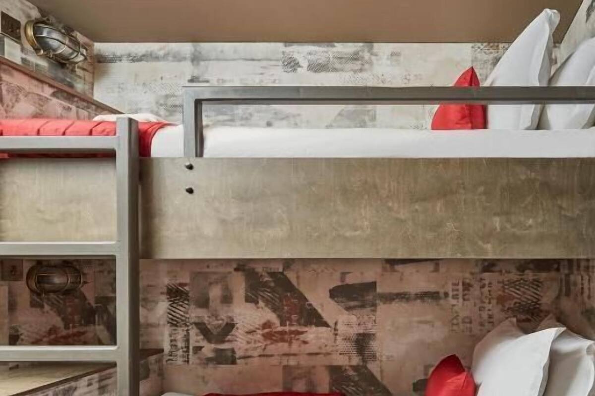 A bunk bed room with a ladder, a red and white bedspread, and a wall with a newspaper print design.