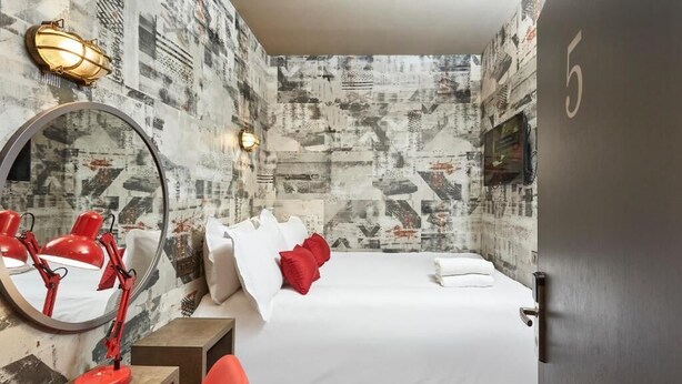 A hotel room with a bed, a desk with a red lamp, a mirror, and a wall with a newspaper print design. - The Pierrooms (Oban)