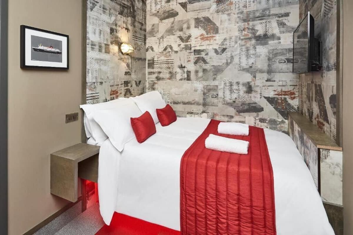 A hotel room with a bed, red pillows, a red blanket, a wall-mounted TV, and a framed picture on the wall.