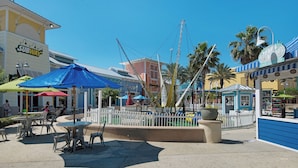 Un espacio exterior soleado con un parque infantil cercado, una sombrilla azul y un restaurante Subway al fondo.