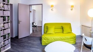 Una habitación luminosa con un sofá verde, una mesa blanca, un televisor sobre un mueble bajo y estanterías.