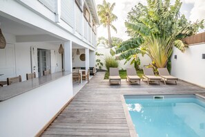 A poolside area with a wooden deck, lounge chairs, and a dining area with wicker chairs and a table.