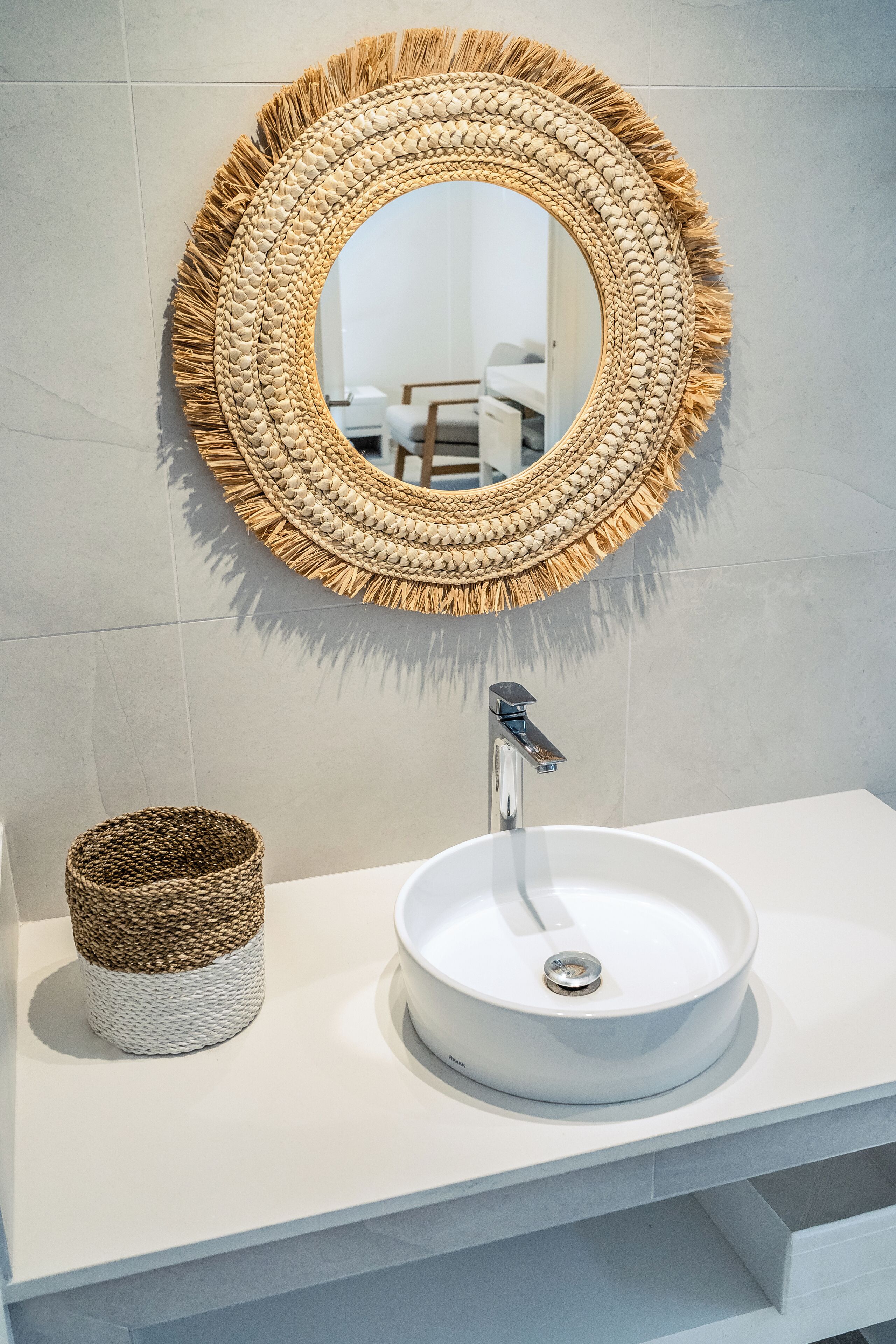 Une salle de bain avec un miroir rond, un lavabo blanc et un panier en osier.