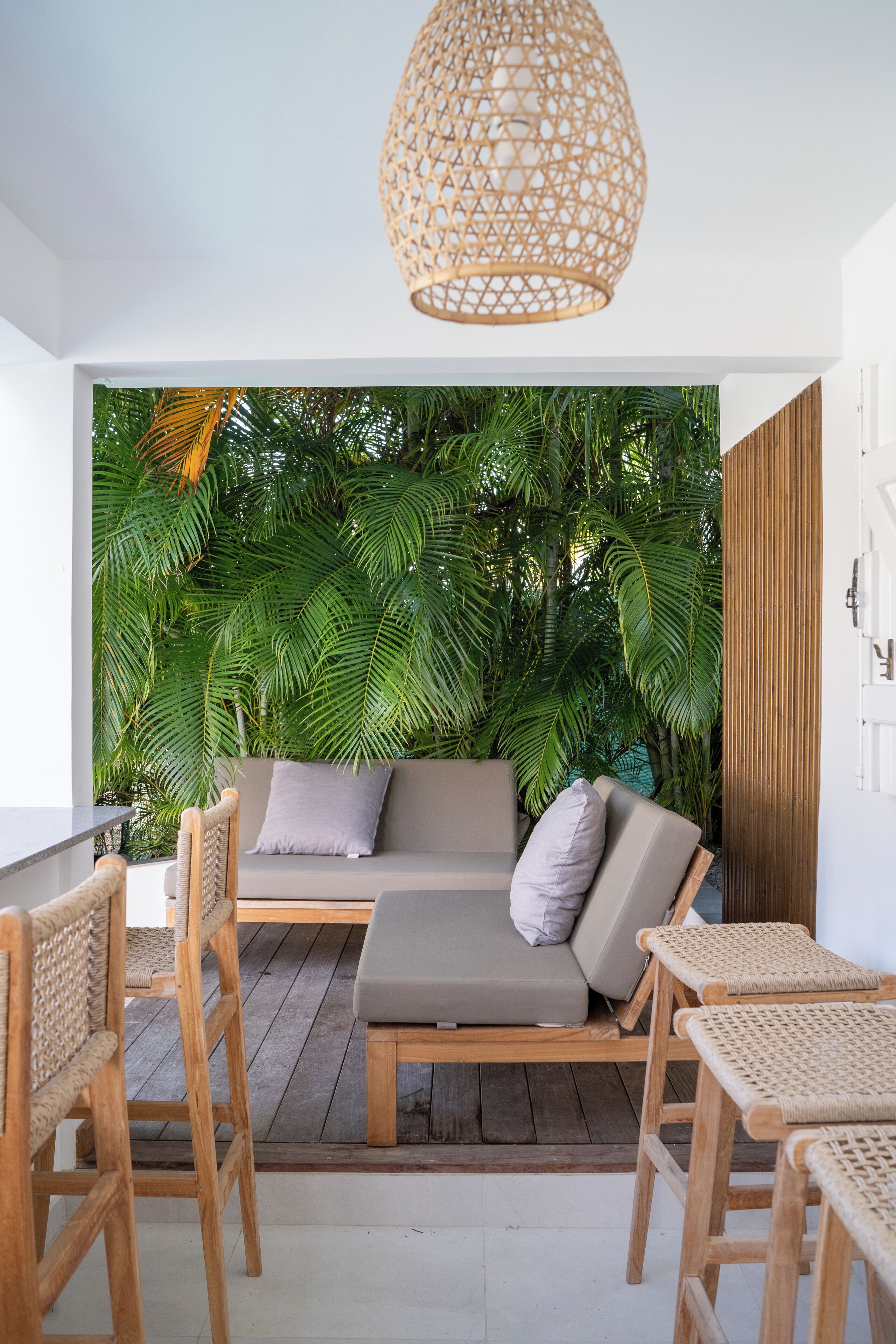 Uma área de estar externa moderna com sofá, cadeiras e mesa, cercada por vegetação exuberante e um grande candeeiro de fibra natural.