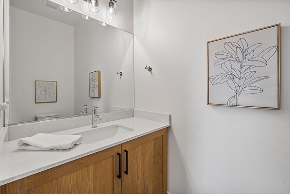 A modern bathroom with a white countertop, wooden cabinets, a large mirror, and a framed botanical print on the wall.