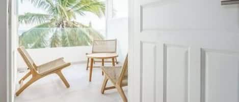 Une pièce avec une porte blanche, une fenêtre donnant sur des palmiers et deux chaises en bois.