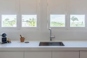 A kitchen with a white countertop, a sink, and three windows with a view of greenery.