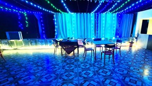 Une scène avec des rideaux bleus et des guirlandes lumineuses, une table avec des chaises et un comptoir où sont servis des plats, le tout illuminé par un néon.