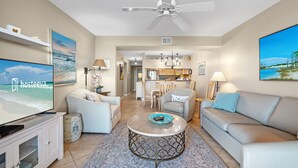 A modern living room with a flat-screen TV, a ceiling fan, a sofa, a coffee table, and a dining area in the background. - Compass Point 508 (Gulf Shores)
