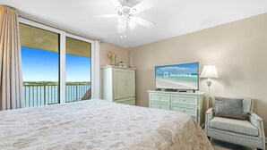 A bedroom with a large bed, a television, a chair, a ceiling fan, and a view of the sea through the window. - Compass Point 508 (Gulf Shores)