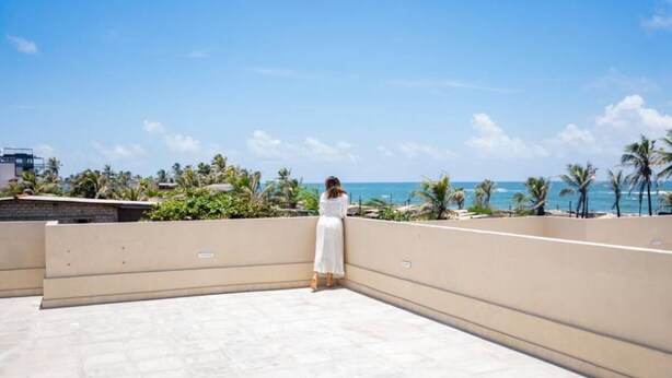 A woman in a white dress standing on a rooftop terrace with a view of the ocean and palm trees.