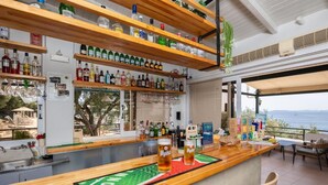 Zona de bar con estantes llenos de botellas de licor, una barra con dos vasos de cerveza y vistas al mar.