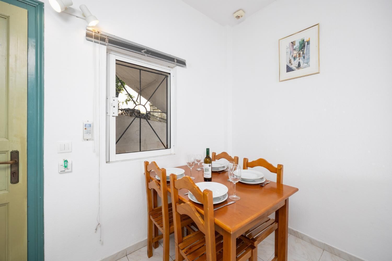 A dining area with a wooden table set for two, a window with a metal grille, and a framed picture on the wall.