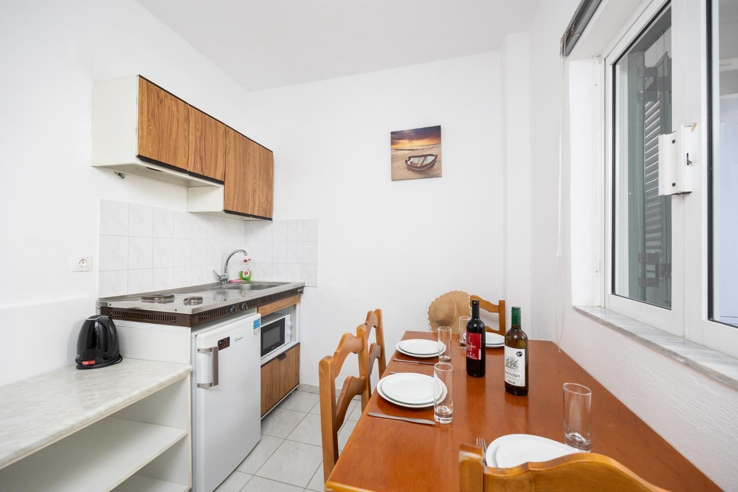 A compact kitchen with a stove, sink, and a small oven. There is a dining table with chairs and a window with blinds.