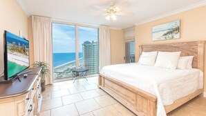 A bedroom with a large bed, a television, a ceiling fan, a view of the beach, and a window with curtains.