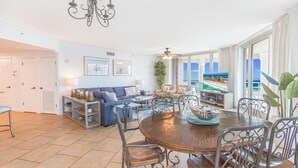 A spacious living area with a large round table, a sofa set, and a television. There is a view of the ocean through large windows.