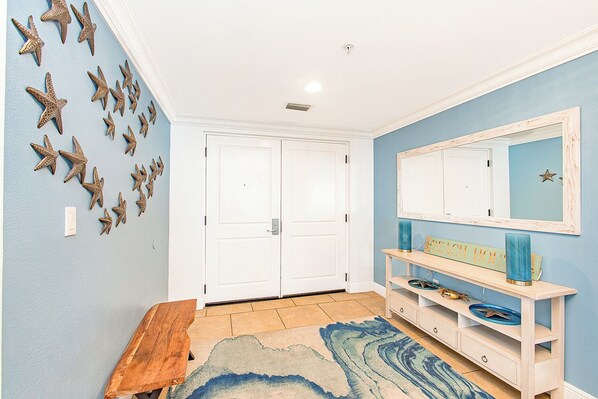 Un pasillo con pared azul, un banco de madera, una puerta blanca y un aparador con espejo y objetos decorativos.
