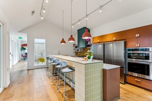 Een moderne keuken met een centraal eiland, barstoelen, roestvrijstalen apparatuur en hangende lampen.