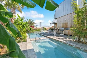 A swimming pool with a wooden deck, surrounded by lush greenery and a modern building in the background. - The Herald at CastleDay - Private Pool, Spa (New Orleans)